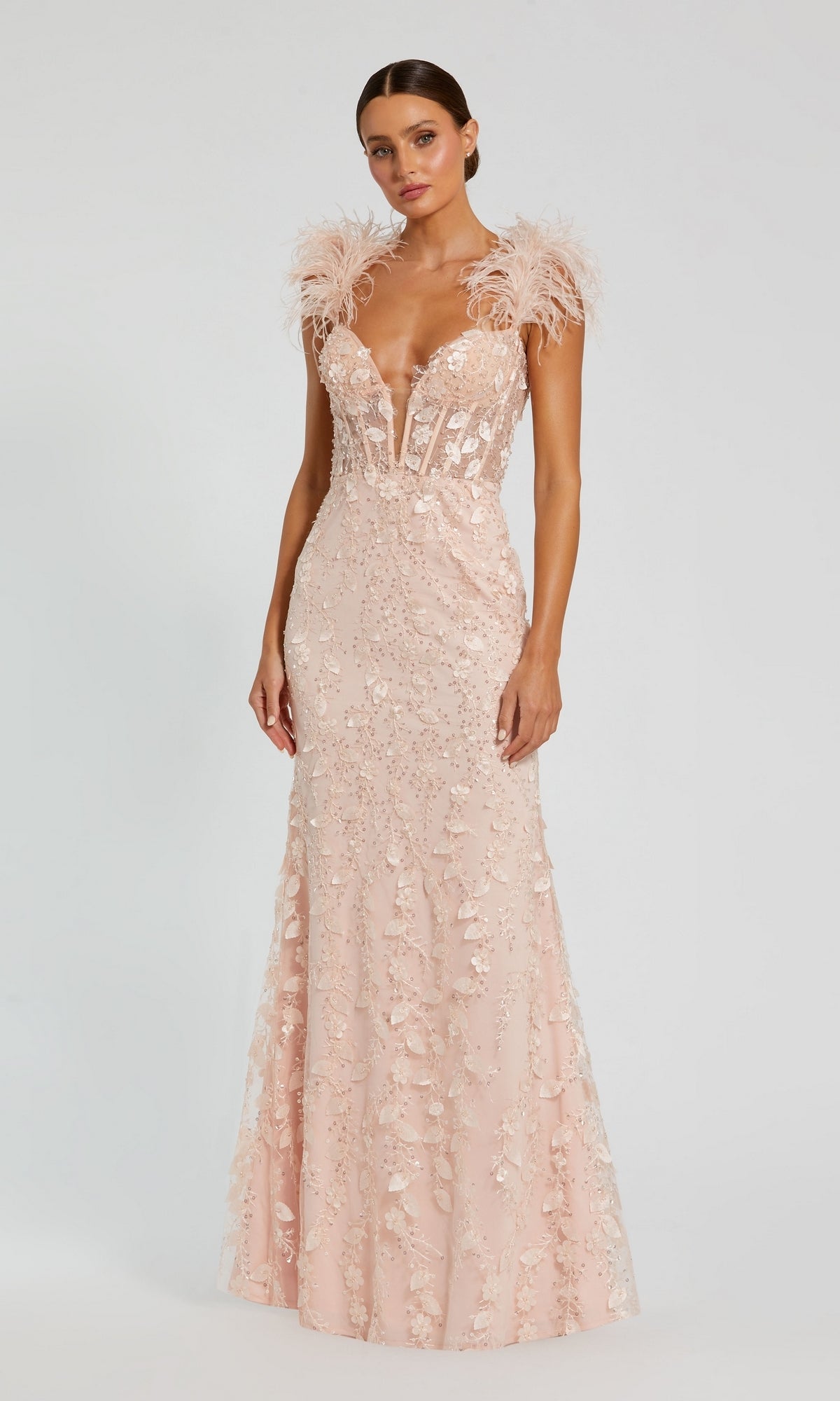 A woman stands against a plain background wearing the Long Formal Dress 20889 by Mac Duggal, a blush pink floor-length gown featuring floral appliqué, a v-neckline, and feather straps.